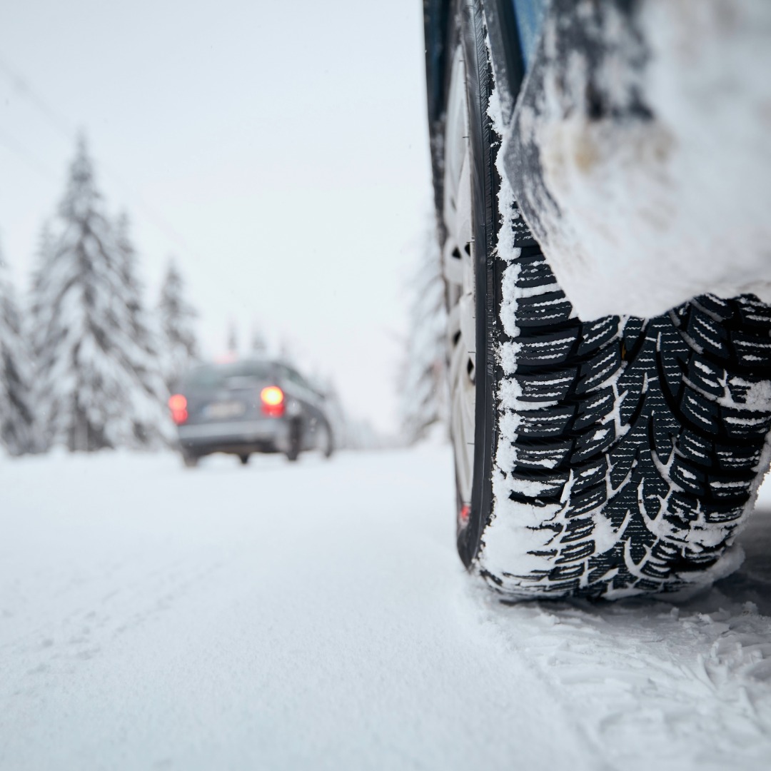 View from the front window of a car, on a grey day, while driving on a highway. It is snowing, the medians are snow-covered, and there is some ice on the windshield.